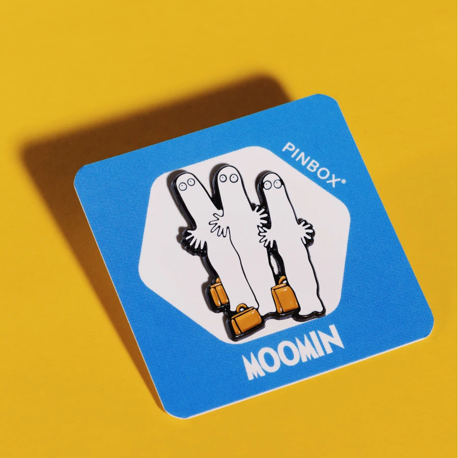Emaliowana pin przedstawiająca trzech Hatifnatów w minimalistycznym czarno-białym designie, trzymających się za ręce z brązowymi walizkami na niebieskiej karteczce z napisem Moomin
