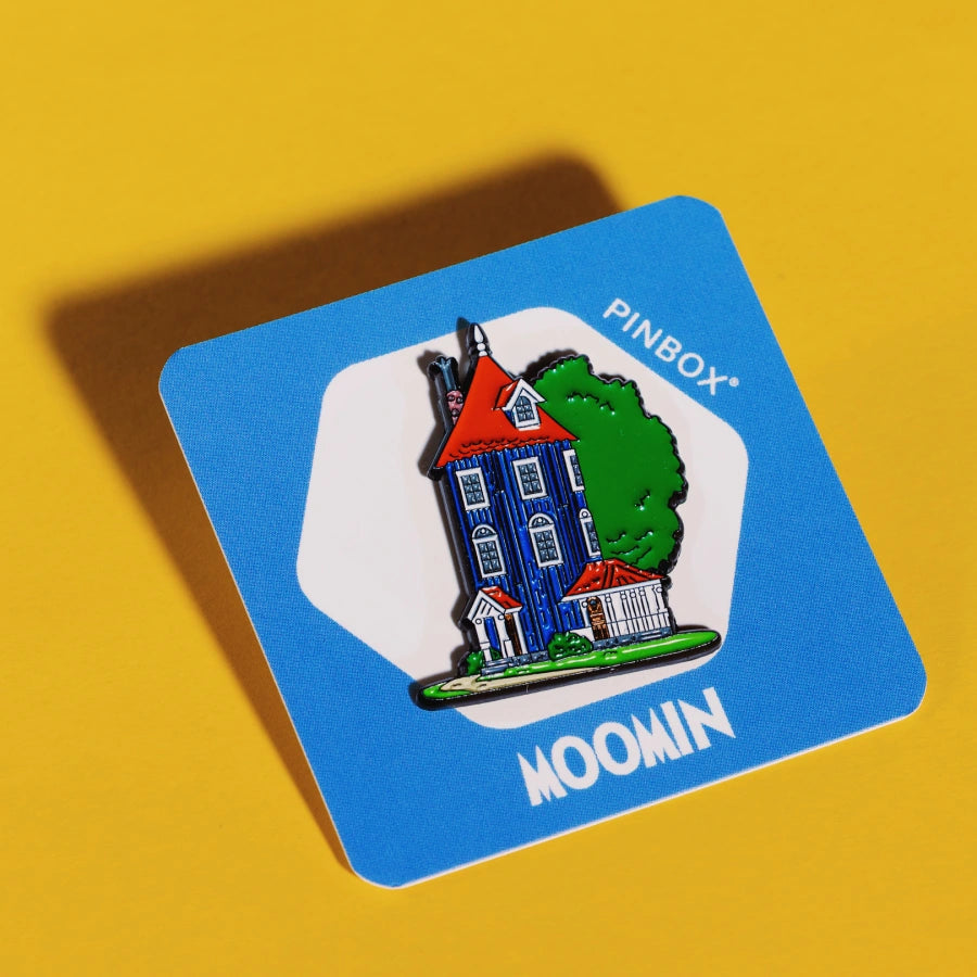 Kolorowy pin przedstawiający Domek Muminków – wysoki niebieski dom z czerwonym dachem i zielonym drzewem, na żółtym tle.